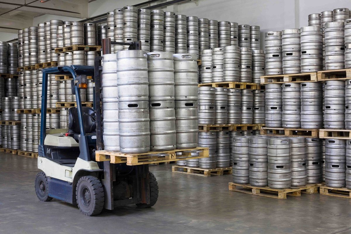 Steel beer kegs stacked on pallets in a warehouse, with a forklift in front